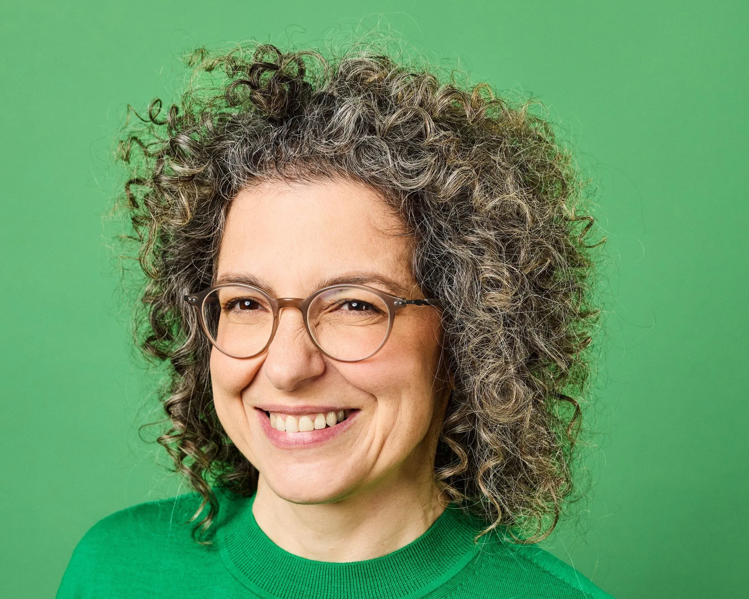 Dr Tzippora Ben-Harim smiling with curly hair and glasses against a green background