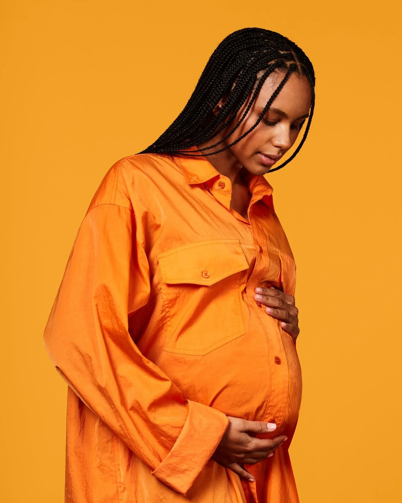 Pregnant woman with long braids in an orange shirt looking down and cradling her belly