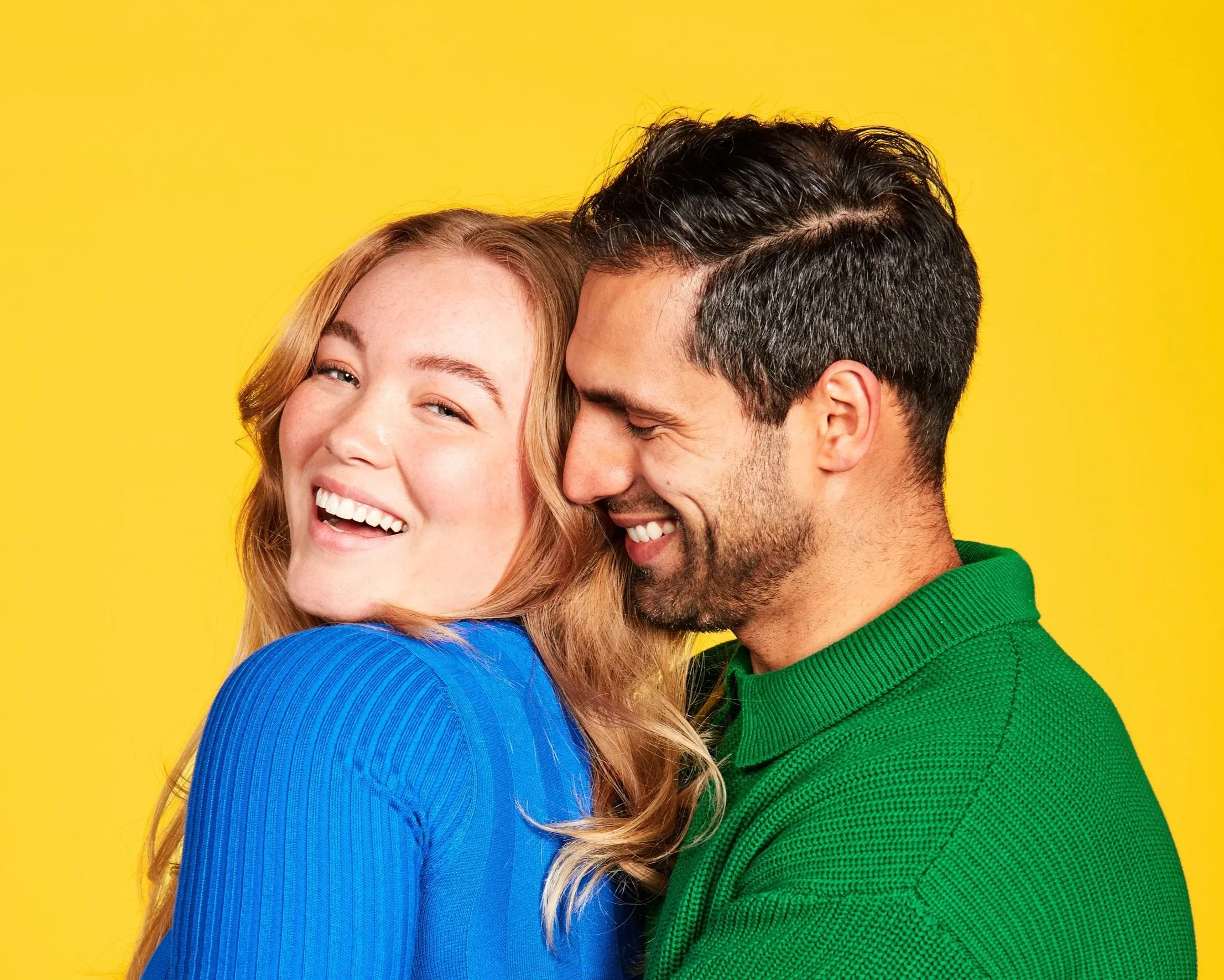 Smiling woman with long blonde hair leaning against a man in a green knit polo, yellow backdrop
