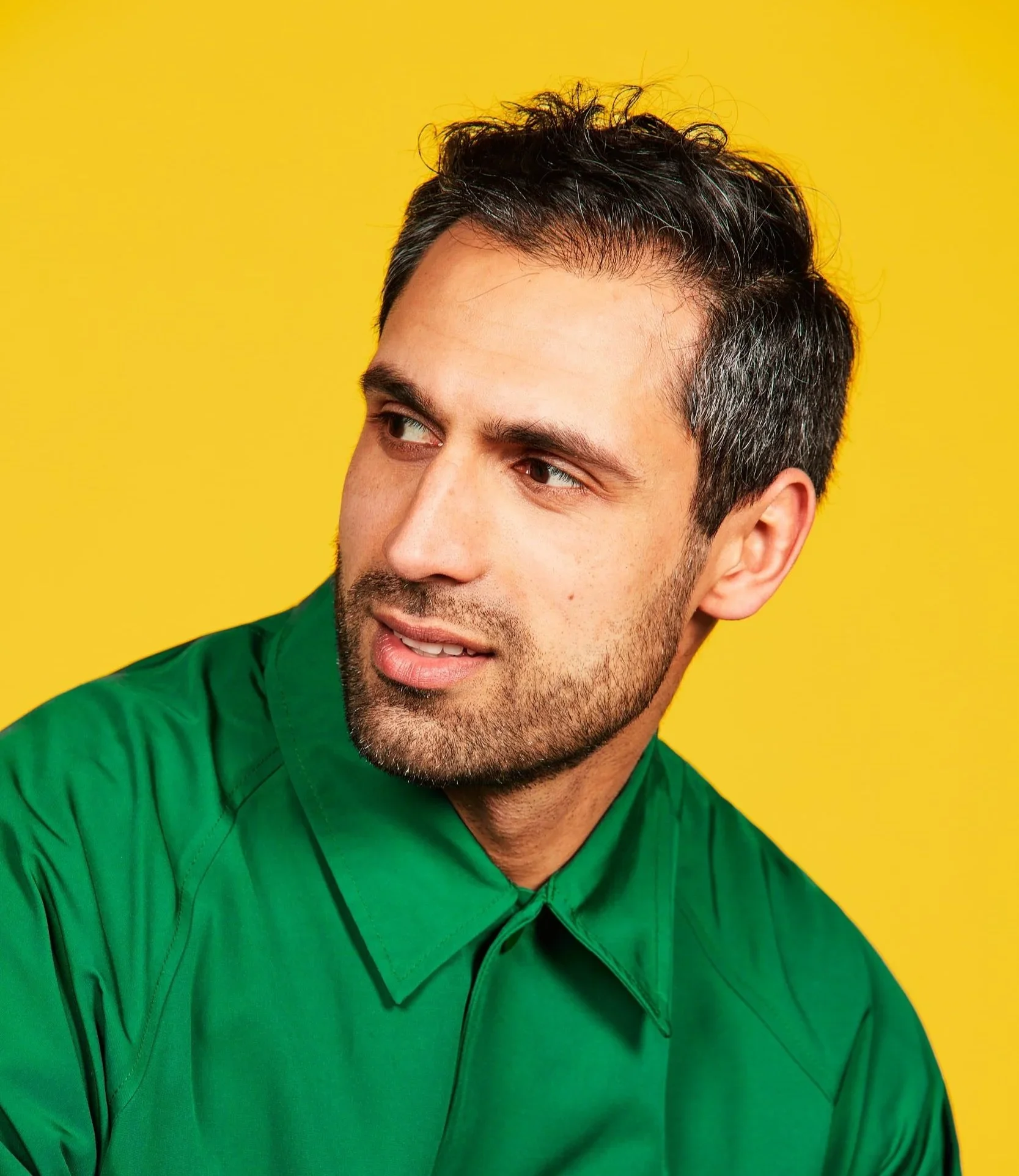 Man with dark hair and stubble in a green shirt glancing to one side against a yellow backdrop