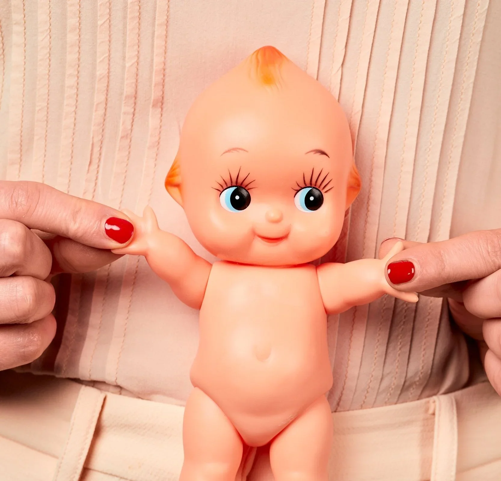Hands holding a vintage Kewpie-style baby doll against a peach pleated blouse