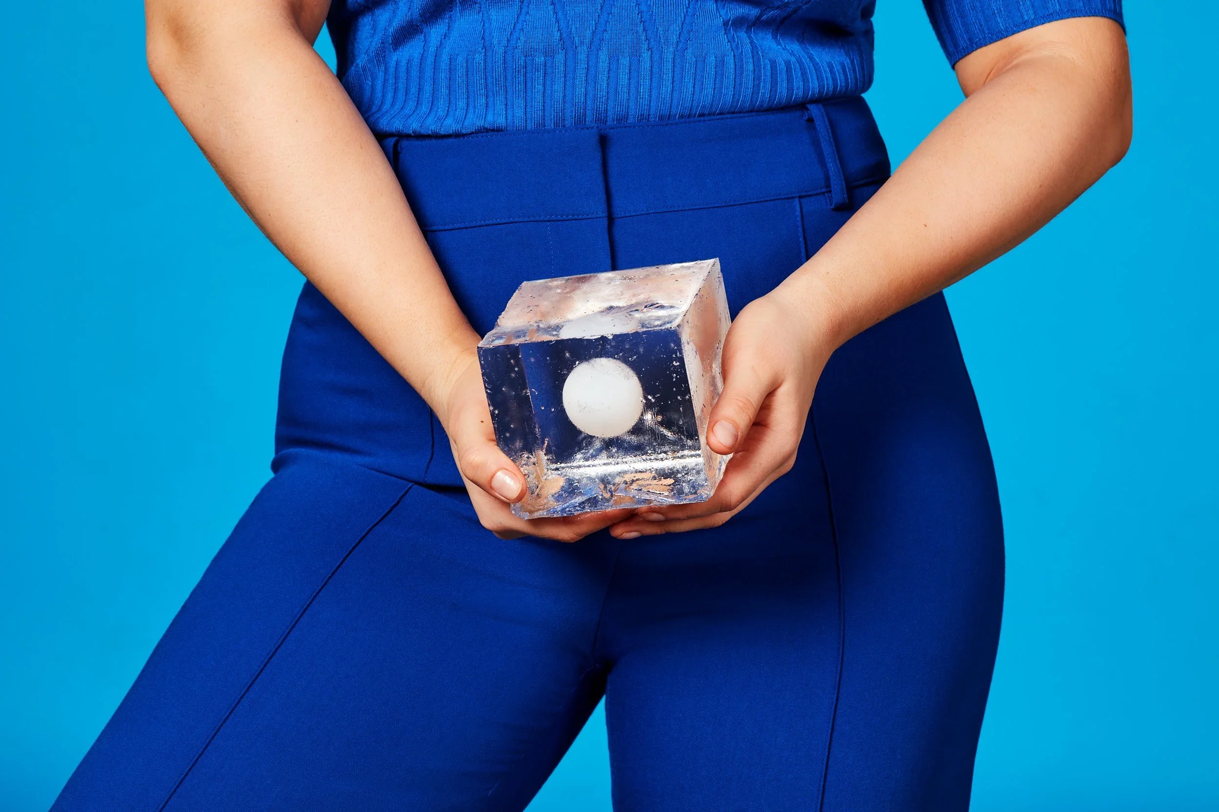 Hands holding a clear ice cube with a single white sphere frozen inside, against a blue backdrop