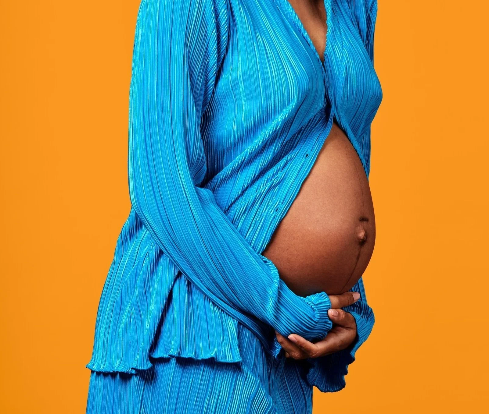 Pregnant woman in a blue pleated shirt cradling her belly against an orange backdrop