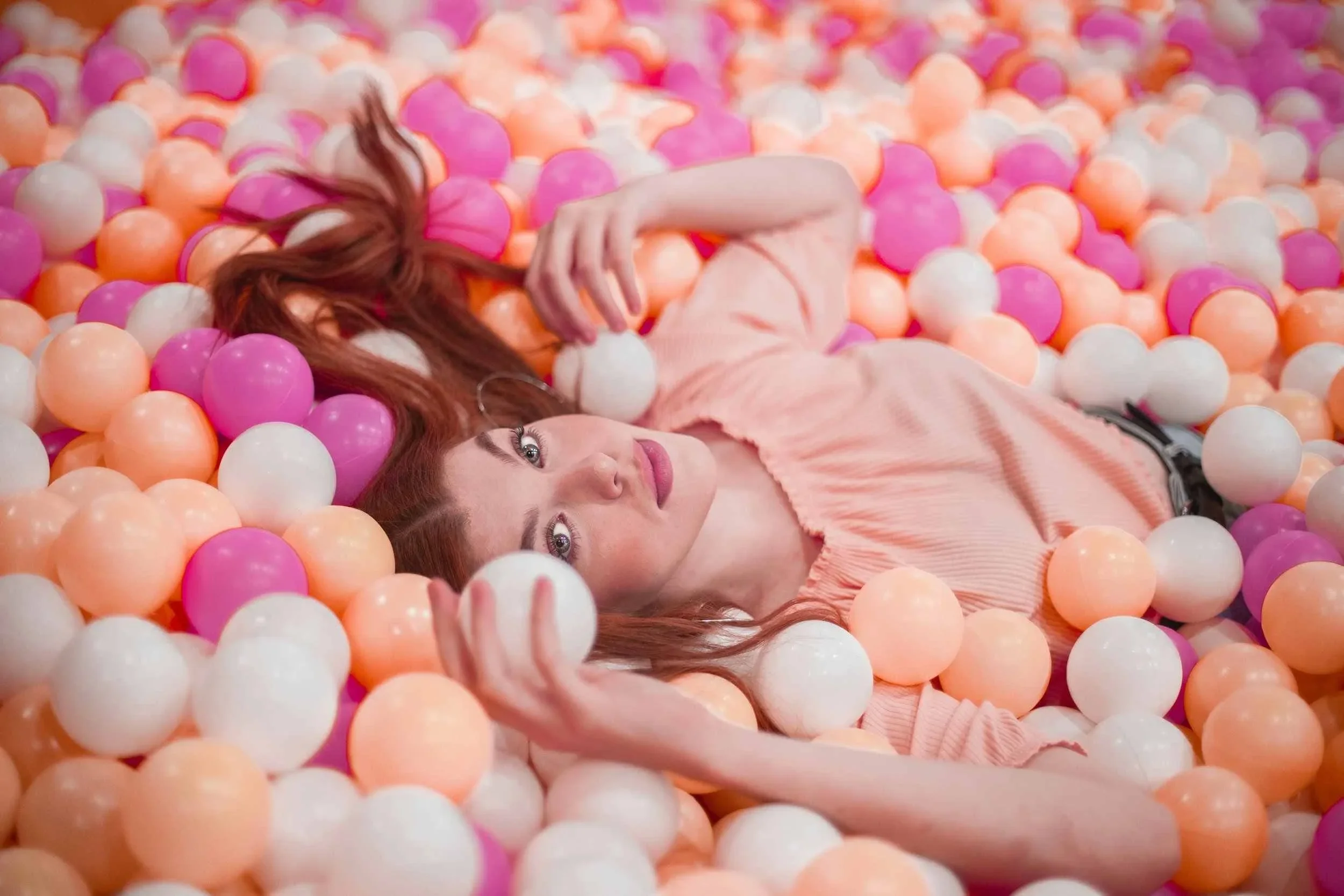 Woman with red hair lying back in a pool of pink, peach and white plastic balls